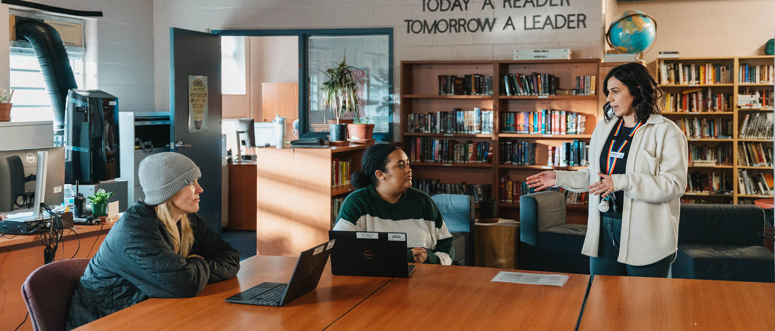 three people talking in a library setting with laptops on a table