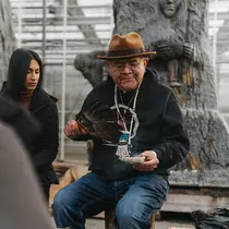 An Indigenous elder performing a traditional ceremony, holding a feather and a bowl, with two people observing attentively in the background.