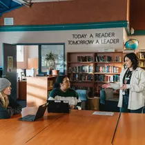 A woman speaking to two others seated at a table in a library, with a sign in the background that reads, "Today a reader, tomorrow a leader."