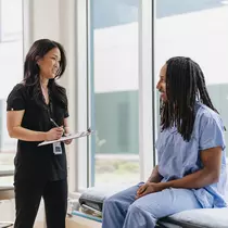 A woman in black scrubs holds a clip board and talks to a man in blue scrubs. 