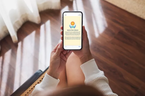A person holding a smartphone displaying a message titled "Advice from Alumni" with a logo and text, viewed from above, in a warmly lit room with wooden floors.