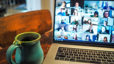 A mug sits on a table next to a laptop. People are in a virtual meeting on the laptop screen.