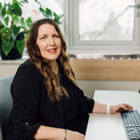 Woman with long brown hairs sits at a desk with a computer keyboard in front of her.