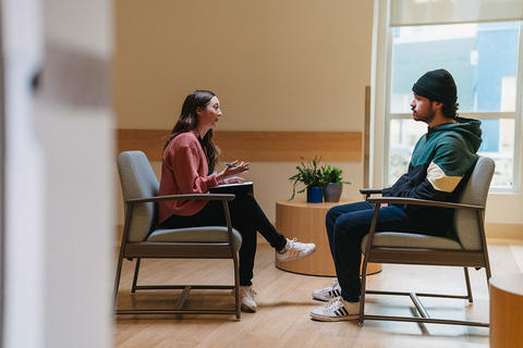 A woman seated on the left and a man seated on the right with a pensive expression. The setting looks like a modern, calm, and well-lit room with a small round table and plants in the background. The interaction seems focused and serious.